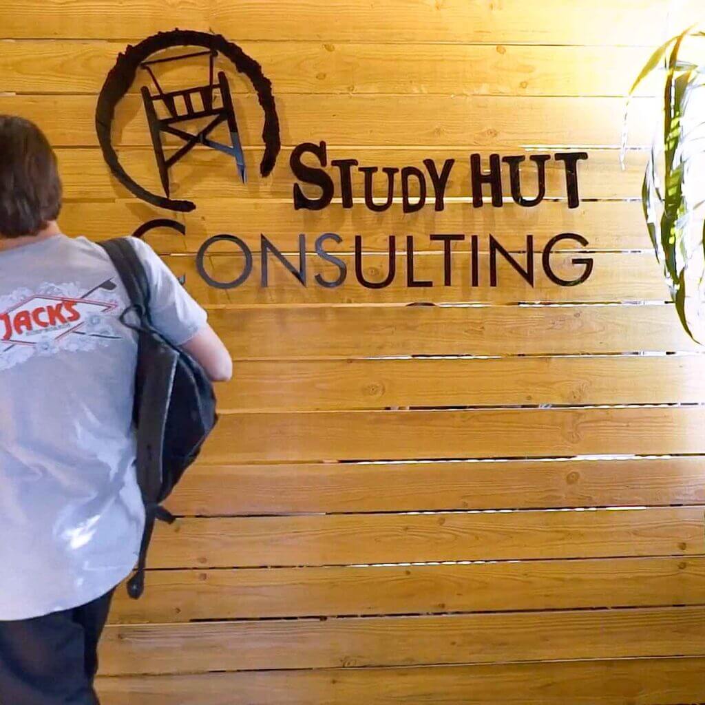 Student walking past a wooden wall with the Study Hut Consulting logo, highlighting a welcoming and professional academic consulting environment.