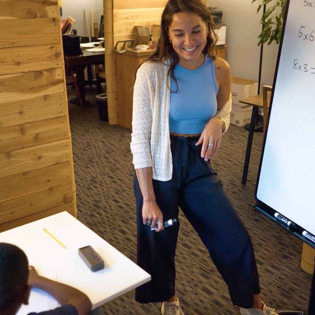 Tutor smiling while helping a young student with math problems at a whiteboard in a cozy, wood-accented Study Hut tutoring center.