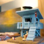 Close-up of a miniature beach scene featuring a blue lifeguard tower, a tiny figurine in swimwear, and a beach ball, set against a blurred background with coastal decor.