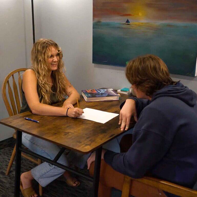 Smiling tutor and student engaged in a relaxed one-on-one session at Study Hut, with books and a sunset painting adding to the welcoming academic atmosphere.
